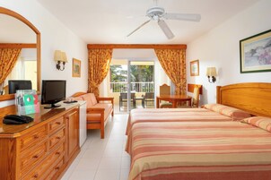 A hotel room with a bed, a desk with a computer, a chair, a TV, and a balcony with a view of palm trees.