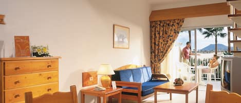 A cozy room with a wooden dresser, a sofa, a table, and a chair. There is a window with a view of palm trees and a mountain.