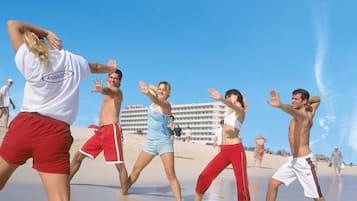 Un grupo de personas bailando en la playa, con un hotel al fondo.