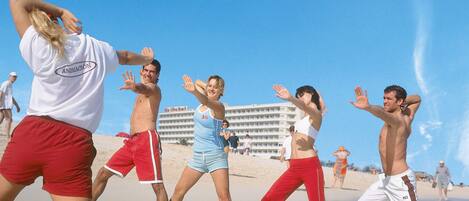 Un grupo de personas bailando en la playa, con un hotel al fondo.