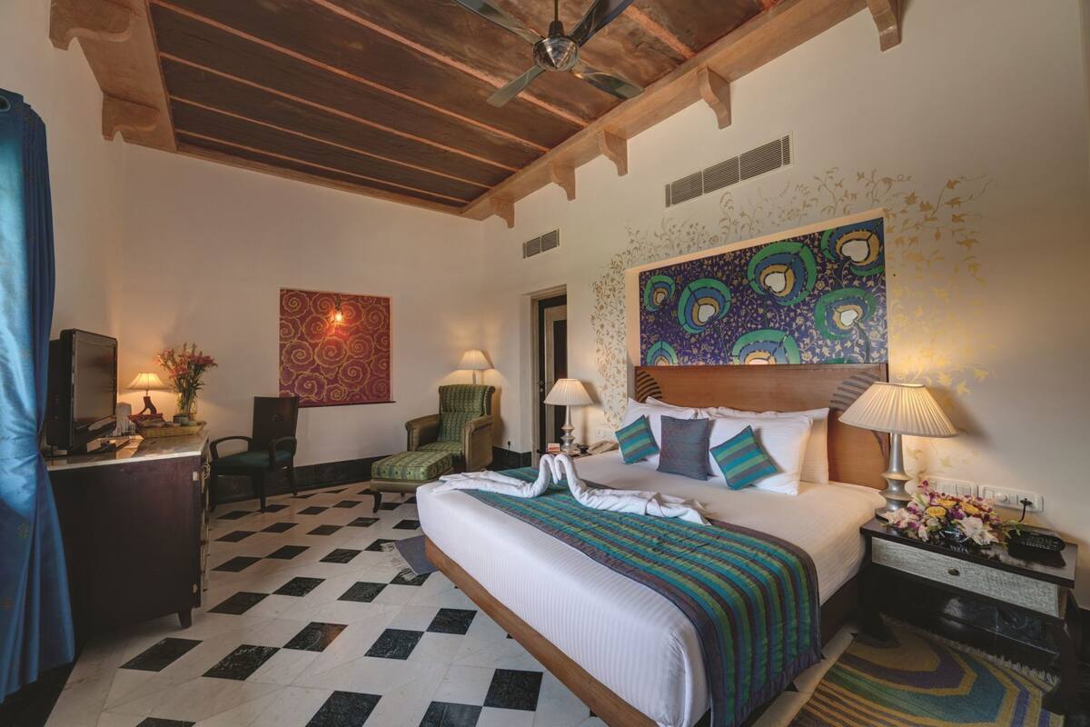 Une chambre d’hôtel avec un sol à carreaux, un lit recouvert d’une couverture à rayures, un bureau avec une lampe, une chaise et un téléviseur.