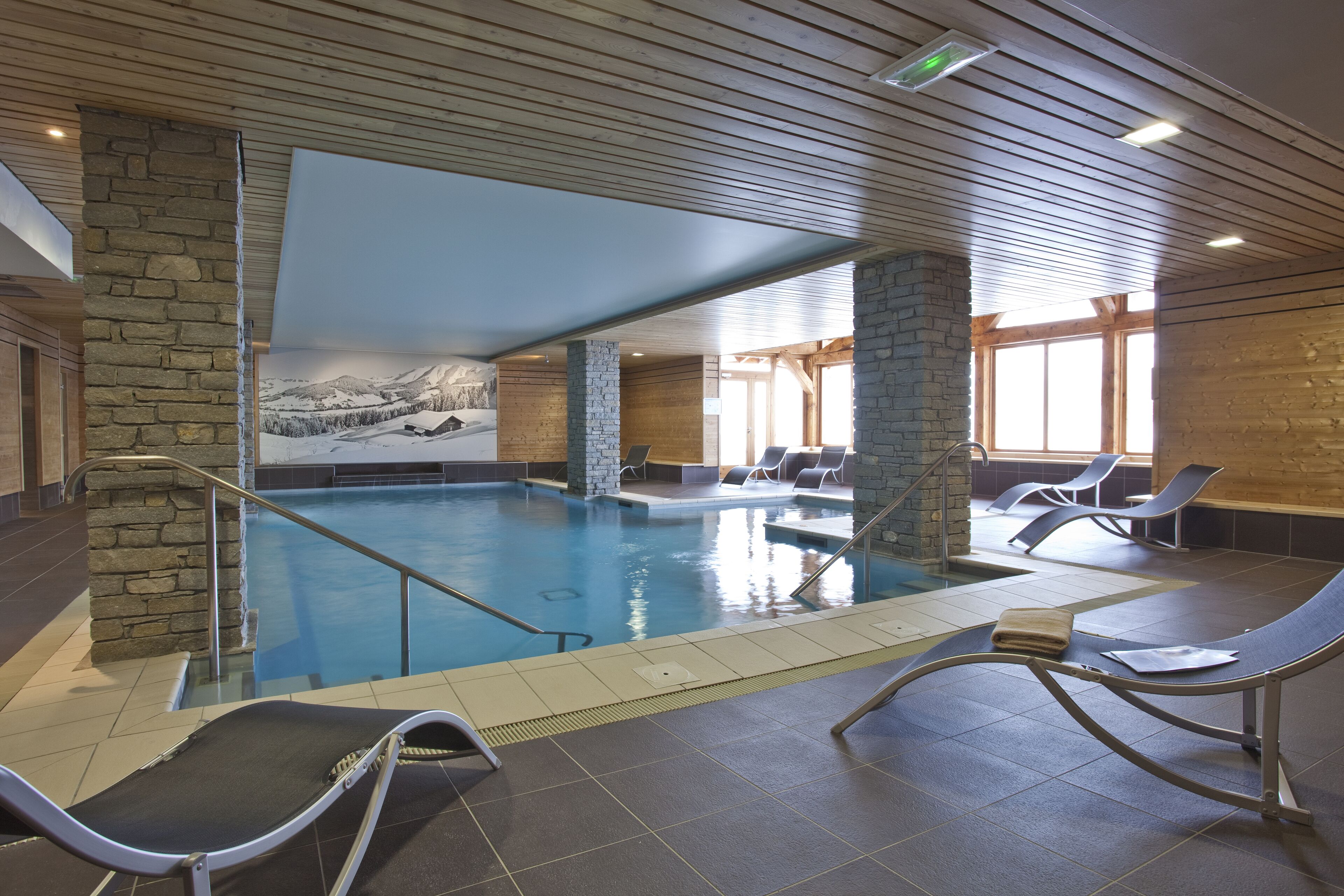 An indoor swimming pool with stone pillars, lounge chairs, and a mural of a snowy landscape.