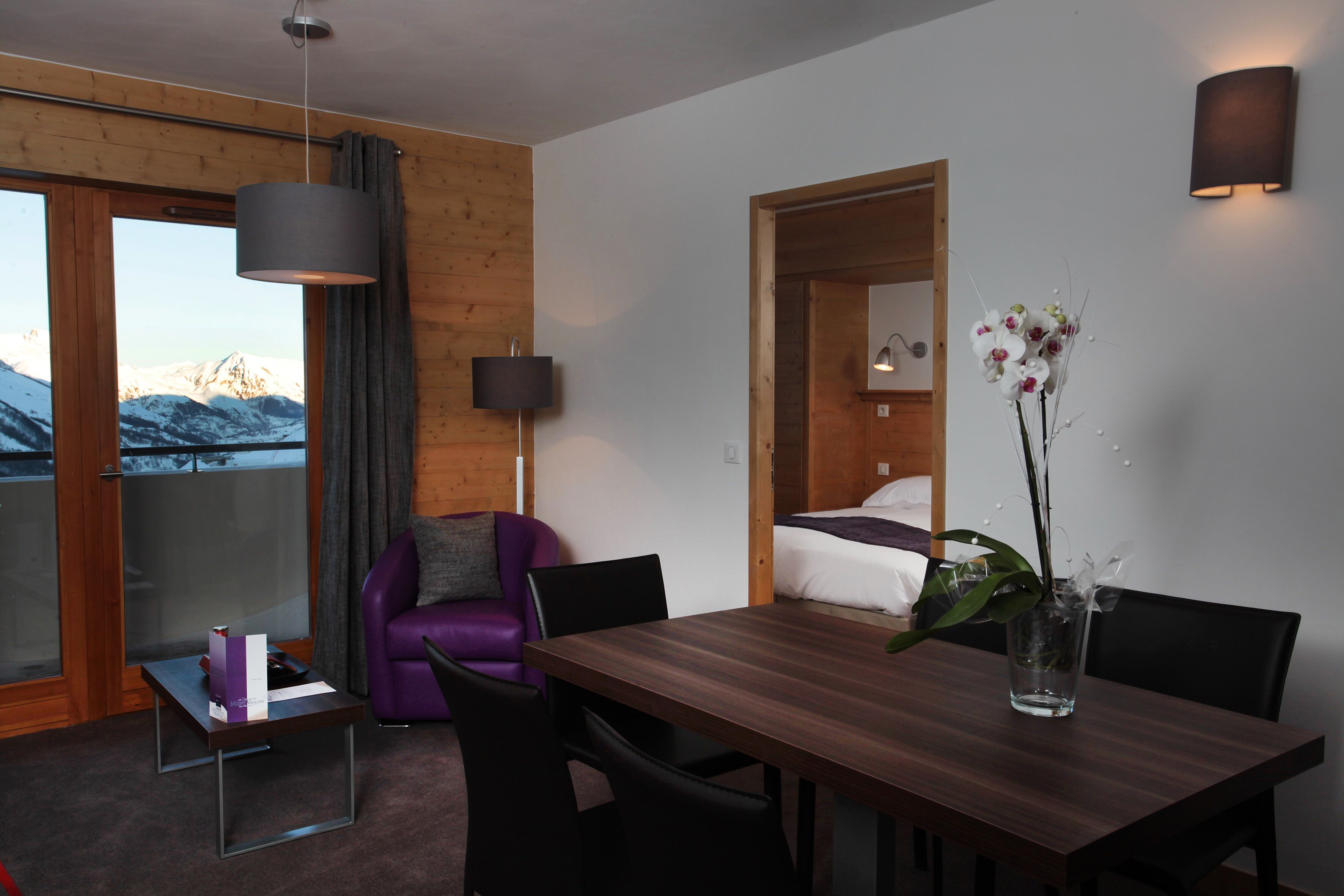 A modern room with a wooden floor, a dining table with chairs, a purple armchair, a small table with a box, a vase with flowers, a bed, a wall-mounted lamp, and a window with a view of mountains.