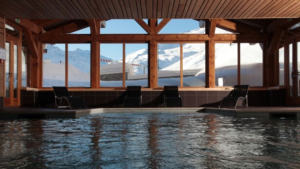 An indoor swimming pool with wooden beams and large windows offering a view of snow-capped mountains. - Le Chalet du Mont Vallon Spa Resort (Les Belleville)