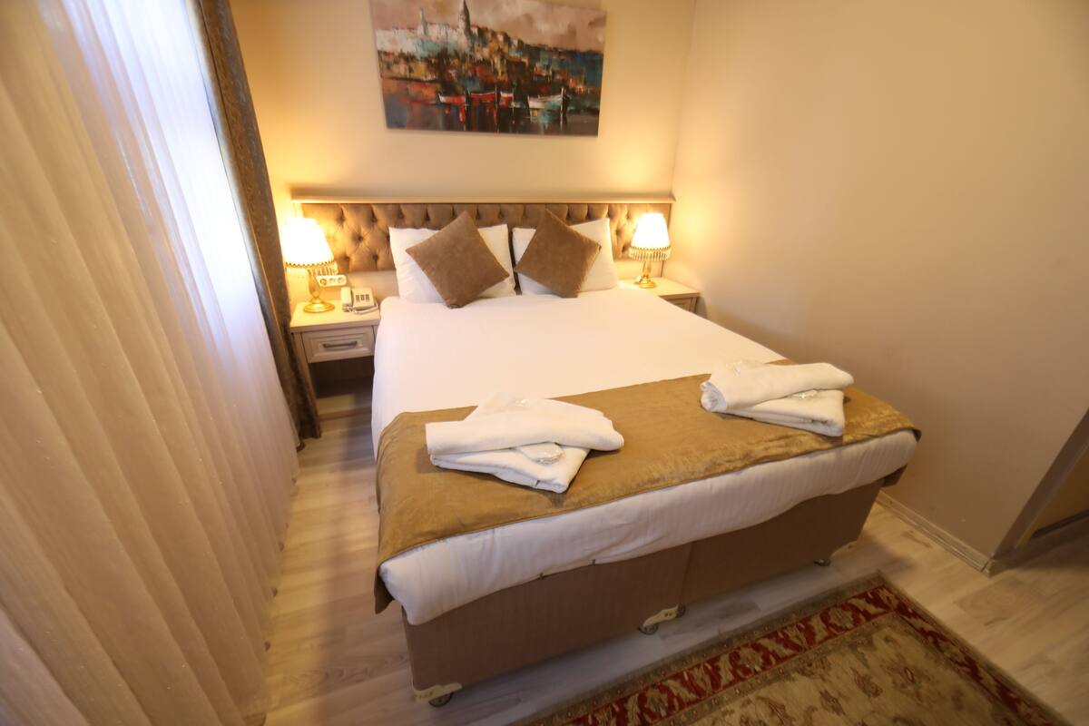 A hotel room with a large bed, two bedside lamps, a nightstand, a painting on the wall, and a patterned rug.