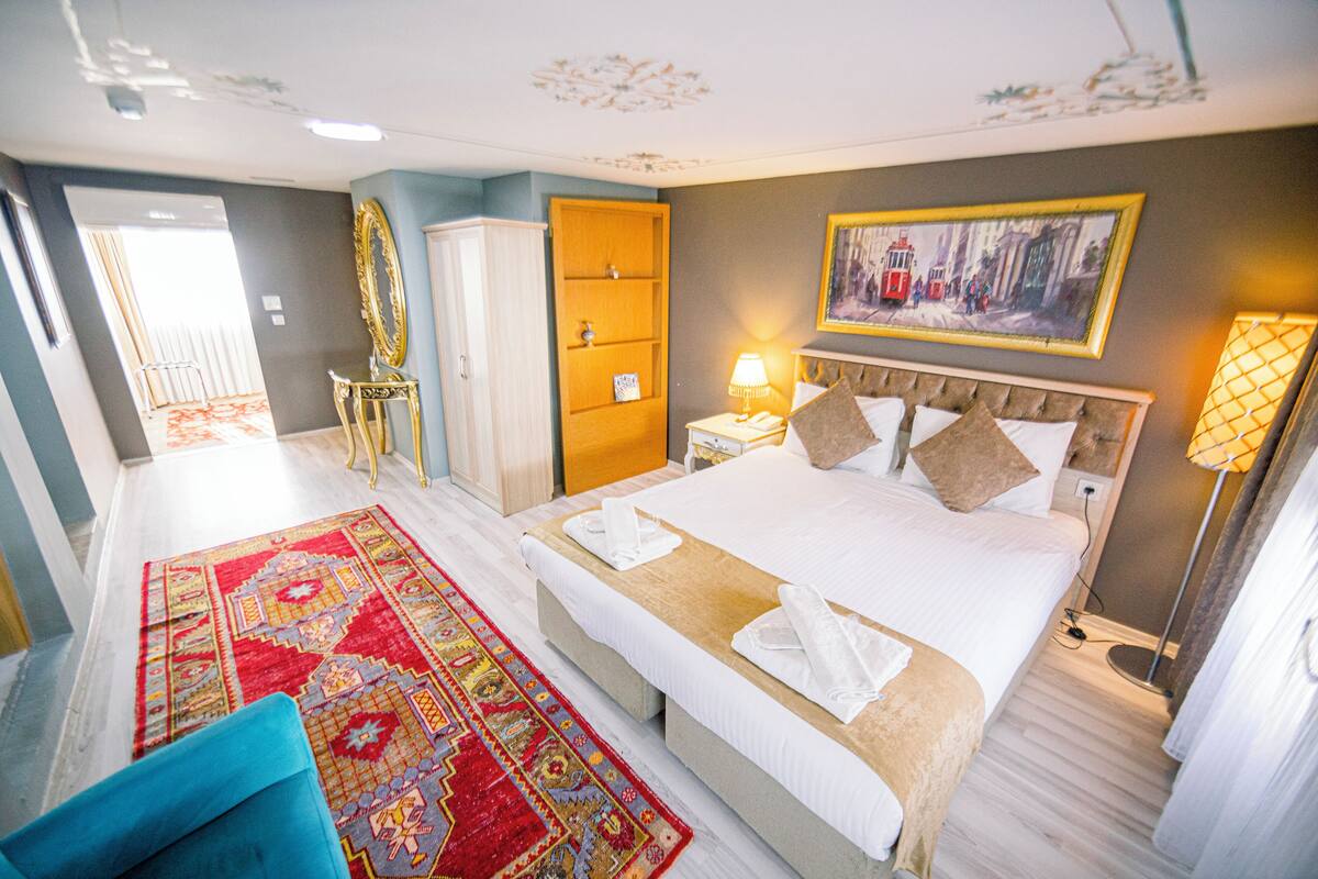 A hotel room with a large bed, a sitting area with a blue chair, a wardrobe, a nightstand with a lamp, and a painting on the wall.