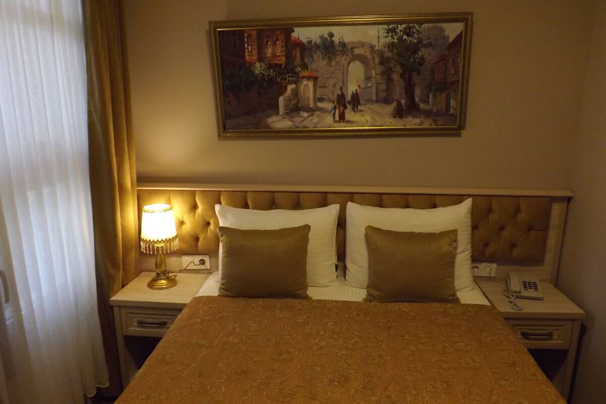 A hotel room with a bed, two bedside tables, a lamp, a painting on the wall, and a curtain.