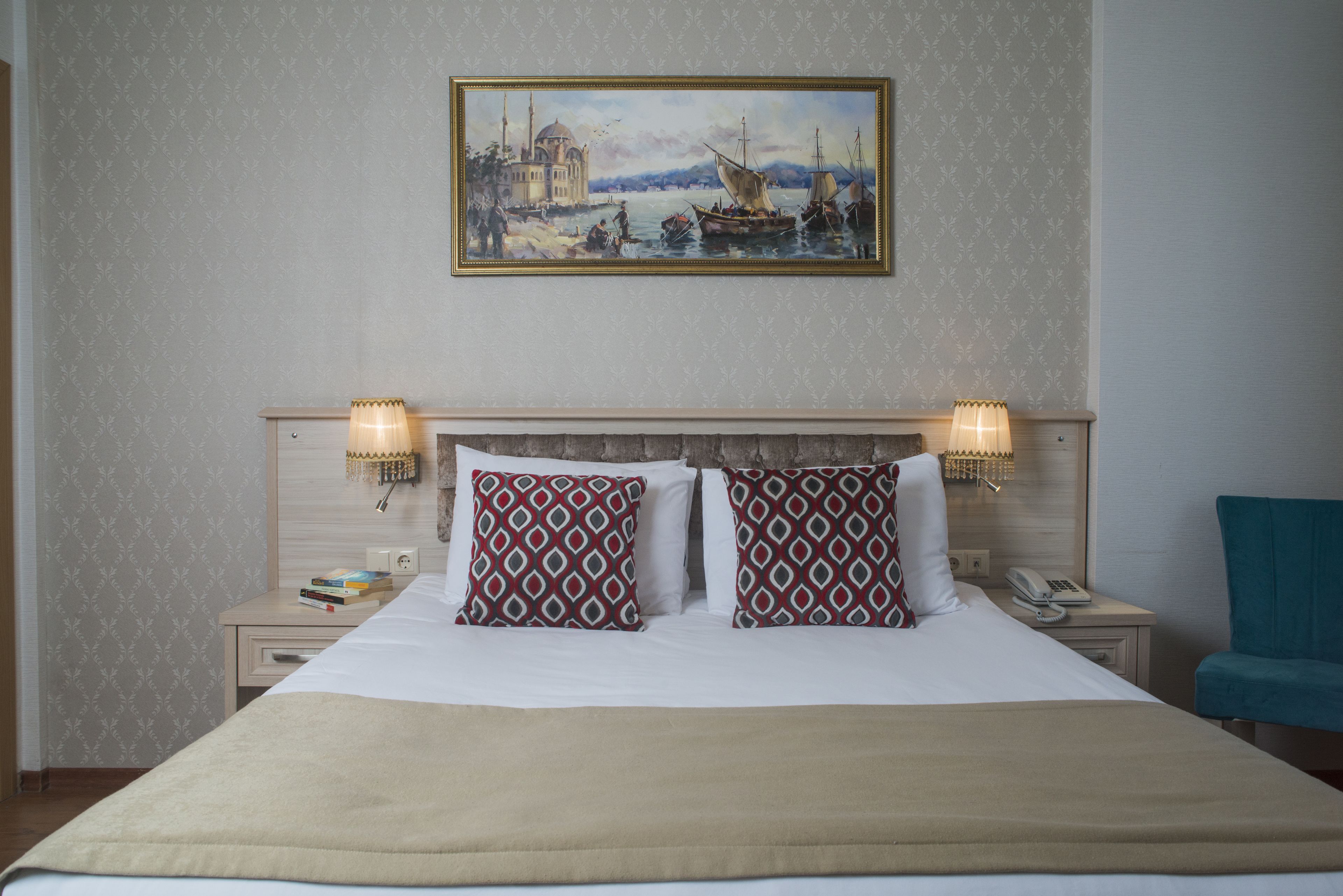 Habitación de hotel con una cama grande, dos almohadas estampadas, una mesita de noche con lámpara, un cuadro de una escena costera y una silla azul.