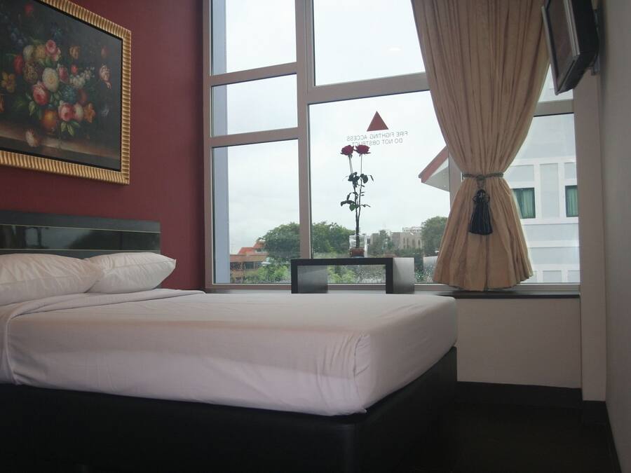 A hotel room with a bed, a painting on the wall, a window with a view of buildings, and a curtain.