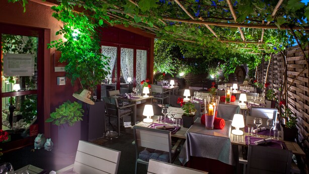 Een terras met tafels klaar voor een maaltijd, omgeven door groen en overdekt met een pergola.