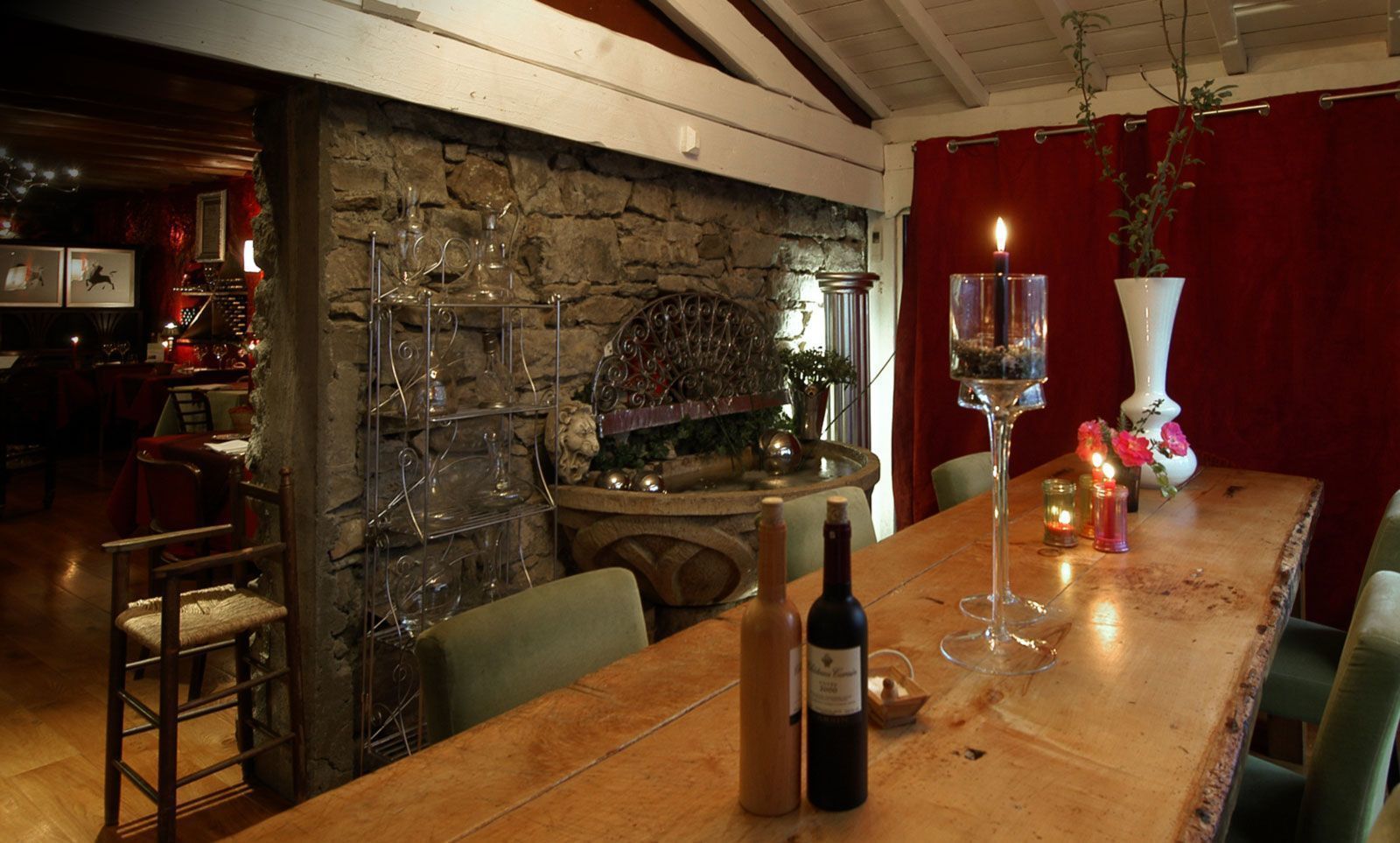 Un coin repas rustique avec un mur en pierre, une table en bois et des chaises. Une bouteille de vin et des verres sont posés sur la table.