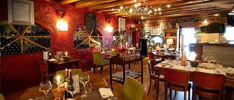 A restaurant with wooden beams, red walls, and a wine rack. Tables are set with wine glasses and candles.