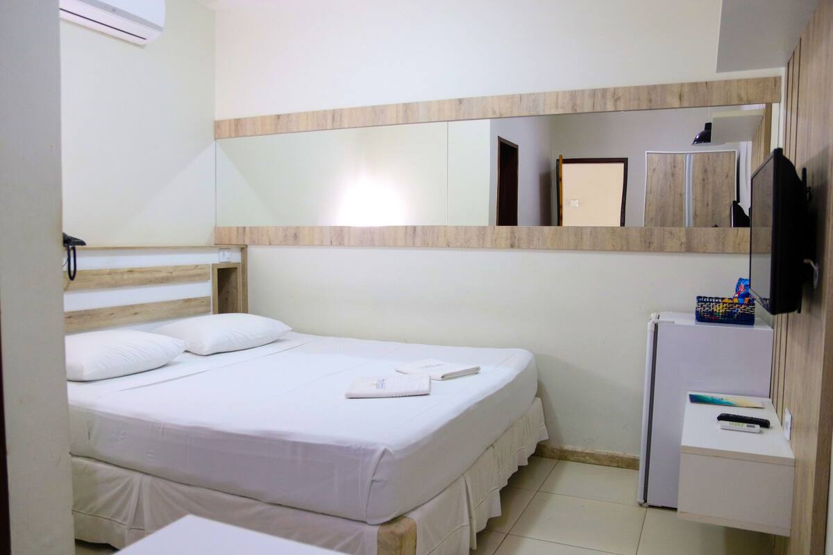 Ein Hotelzimmer mit Bett, kleinem Schreibtisch mit Kühlschrank, Spiegel und Wandfernseher.