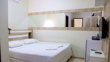A hotel room with a bed, a small desk with a refrigerator, a mirror, and a TV mounted on the wall.