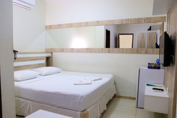 A hotel room with a bed, a small desk with a refrigerator, a mirror, and a TV mounted on the wall. - Imperial Palace Hotel (Barbalha)
