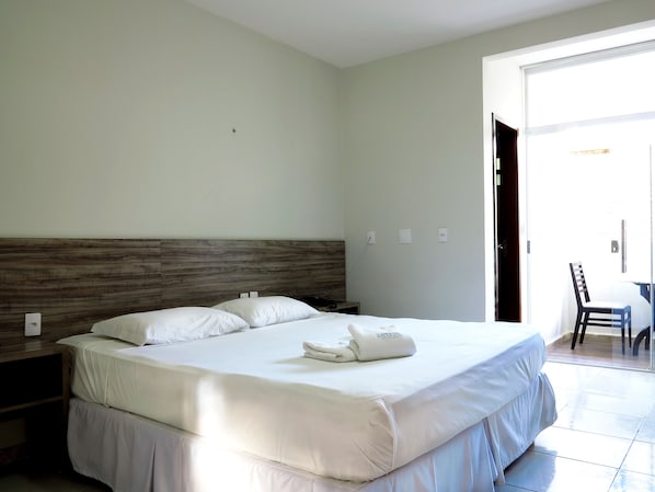 Habitación de hotel con una cama grande, cabecera de madera, mesita de noche pequeña y una silla cerca de la puerta.