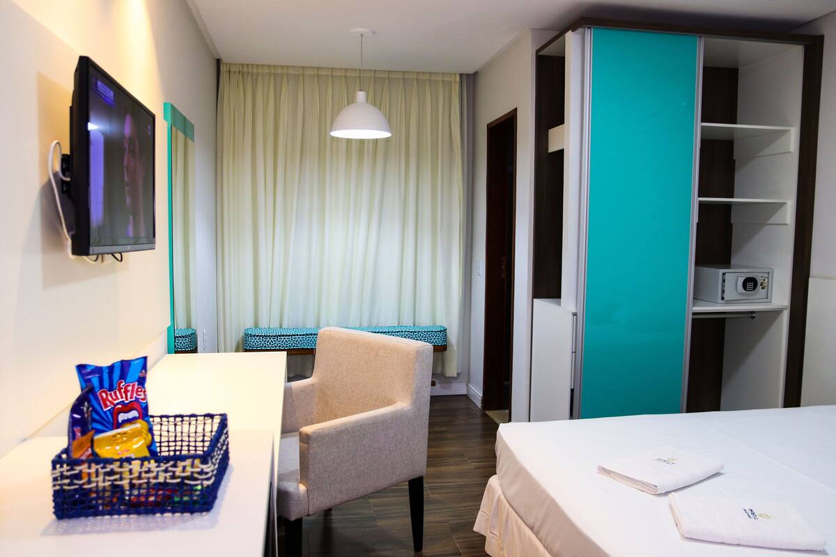 Ein Hotelzimmer mit Bett, Schreibtisch mit Stuhl, Fernseher, Fenster mit Vorhängen und einer Minibar.