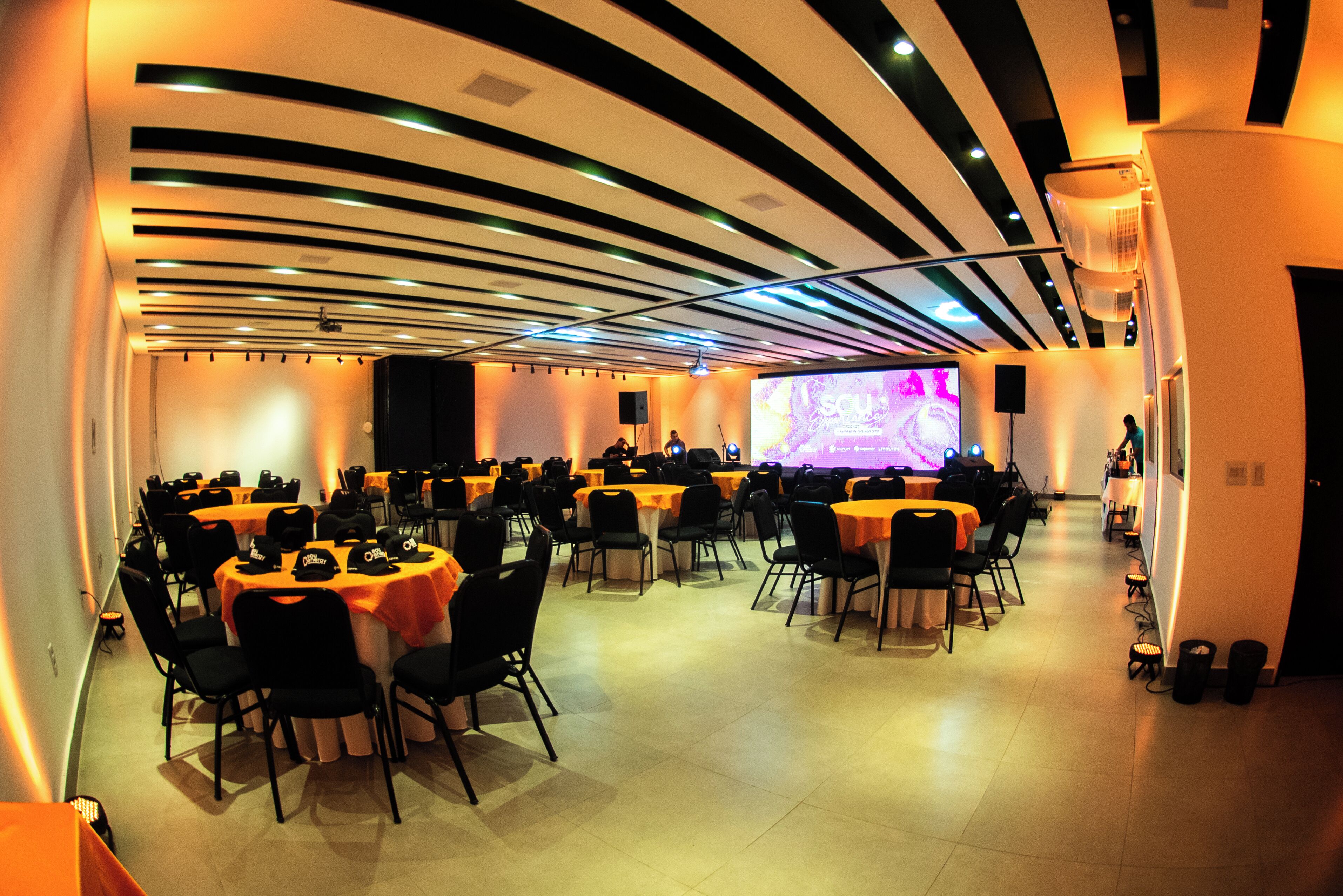 Un salón de conferencias con varias mesas y sillas, una pantalla grande y un escenario con oradores.