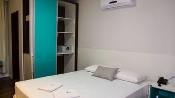 Una habitación de hotel con una cama, una mesita de noche con un teléfono, un estante con un libro y un equipo de aire acondicionado en la pared.
