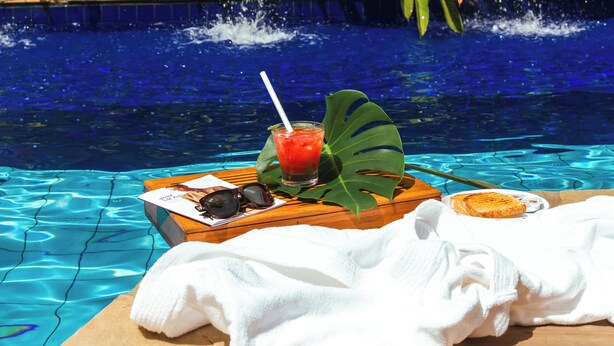 Un área de piscina con una cascada, una mesa de madera con una bebida, gafas de sol y una toalla.