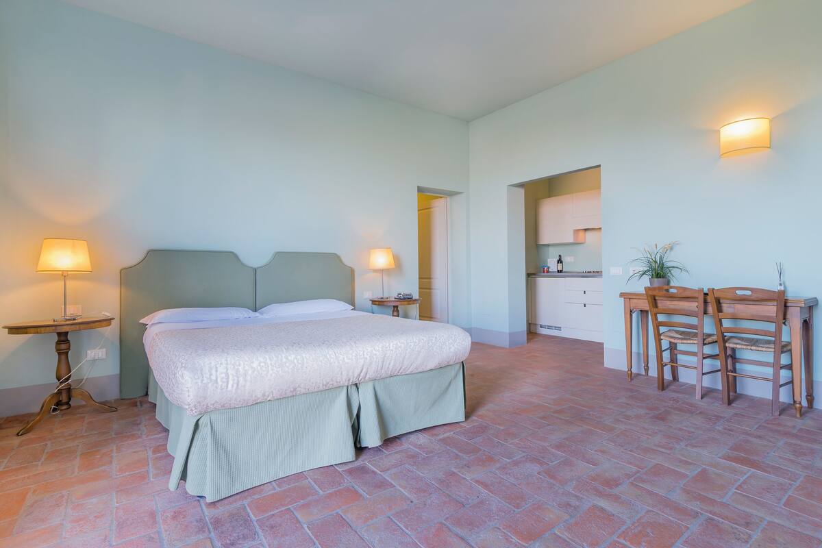 A bedroom with a bed, bedside tables, a desk with chairs, and a kitchen area in the background.