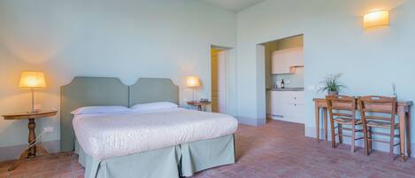 A bedroom with a bed, bedside tables, a desk with chairs, and a kitchen area in the background.