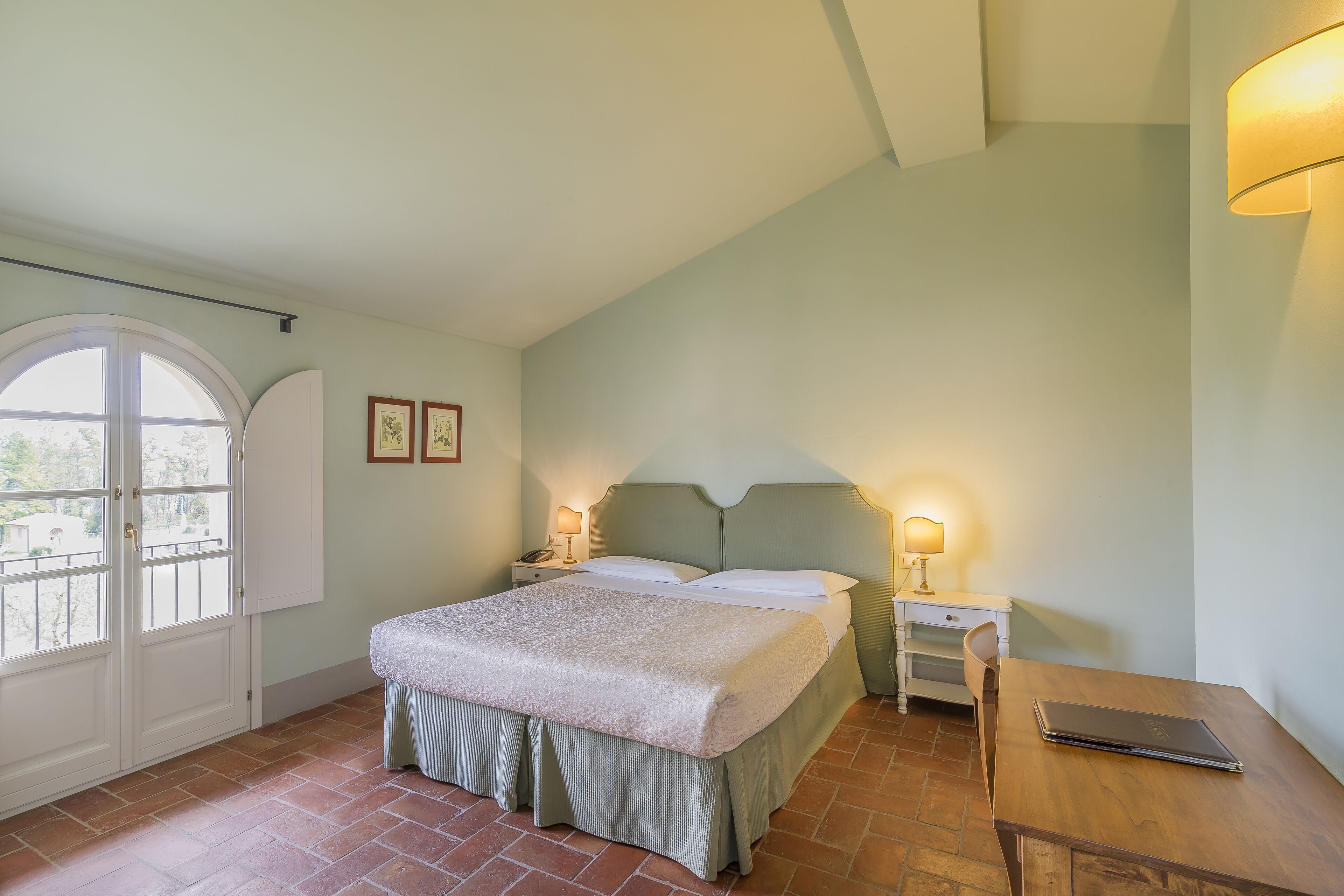Quarto com cama, mesas de cabeceira, escrivaninha e janela.