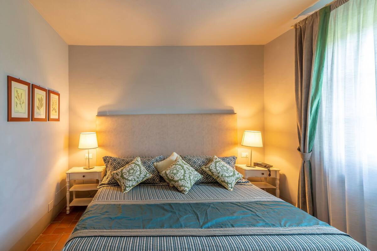 A neatly made bed with patterned pillows and two bedside lamps, a wall-mounted shelf above, and a window with curtains.