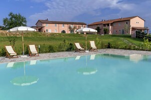 Una piscina con sillas de descanso y sombrillas, rodeada por un jardín ajardinado y un edificio grande al fondo.