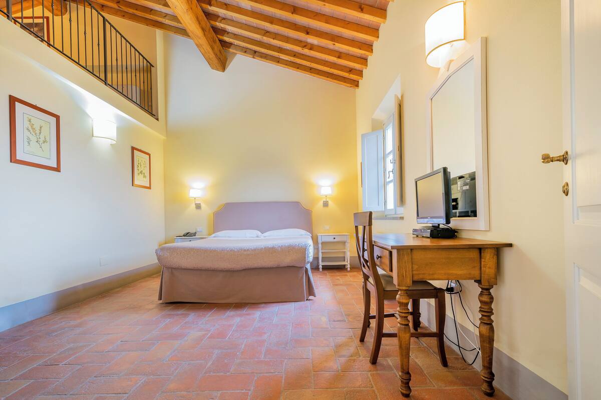 A bedroom with a bed, a desk with a computer, a chair, a TV, and a staircase.