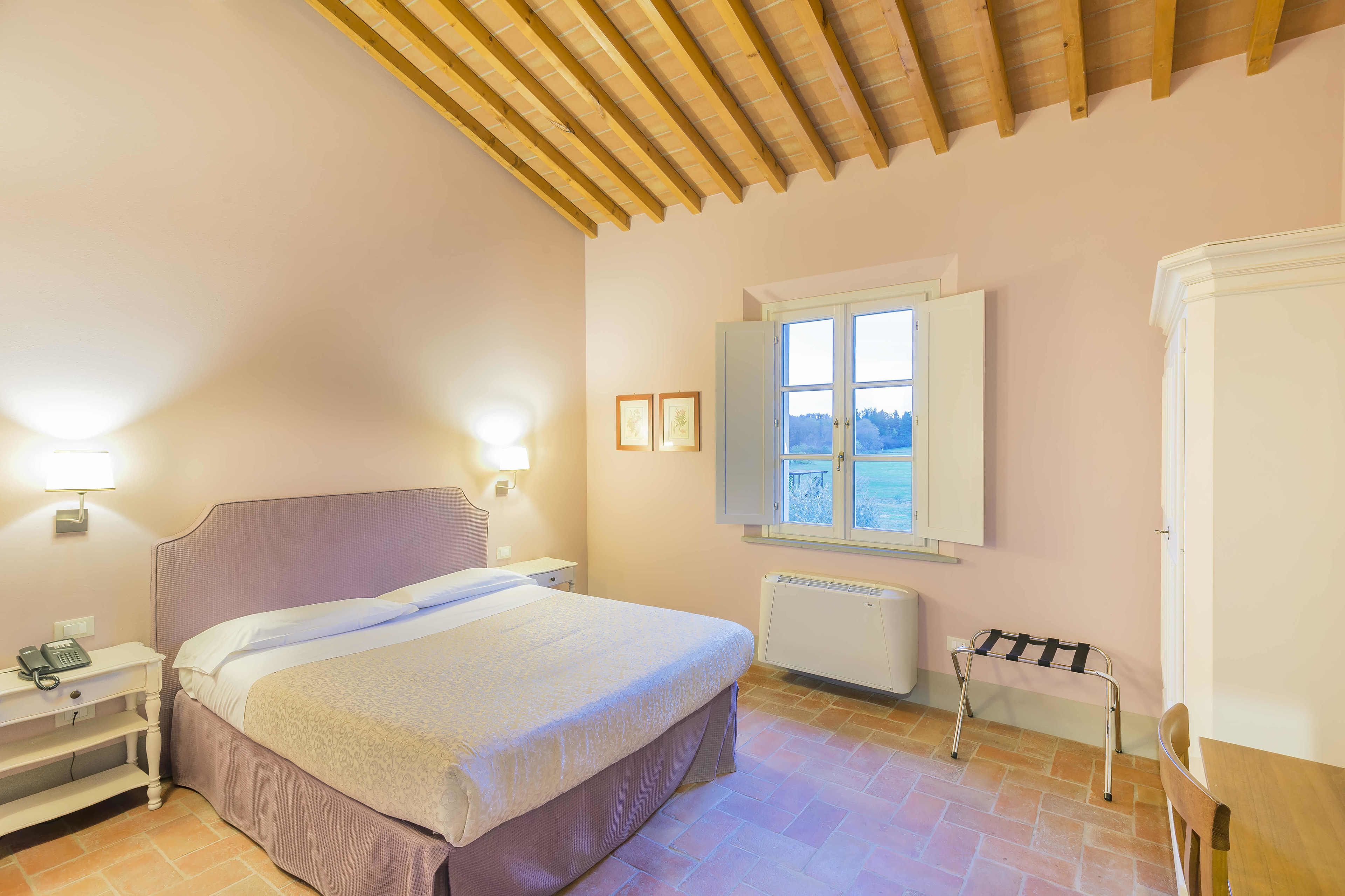 Quarto com cama, mesa de cabeceira com telefone, janela com vista, radiador e teto de madeira.