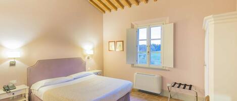 A bedroom with a bed, bedside table with a phone, a window with a view, a radiator, and a wooden ceiling.
