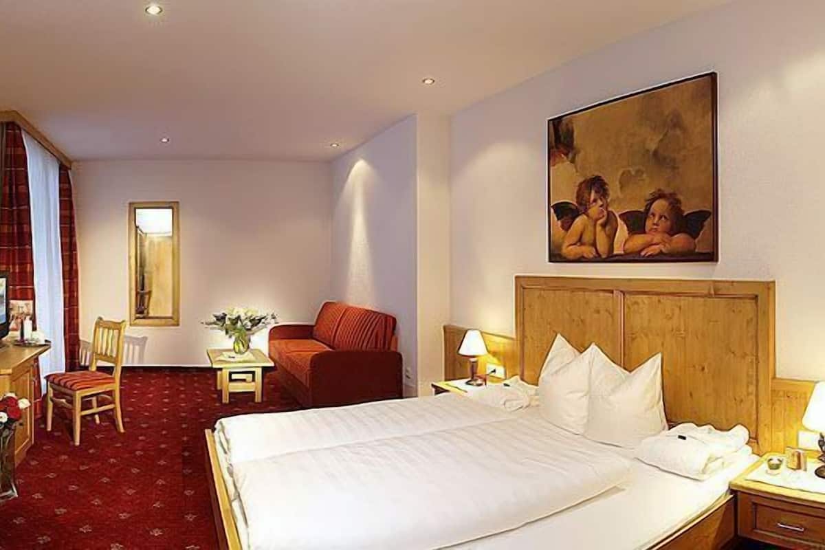 A hotel room with a bed, a desk with a computer, a chair, a small table with a vase of flowers, and a painting on the wall.