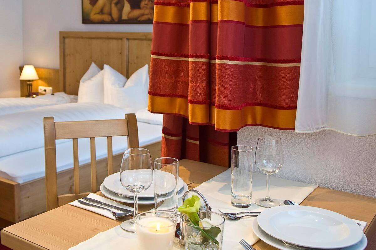 A hotel room with a neatly made bed, a dining table set with plates, glasses, and a candle, and a wooden chair.