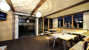 A restaurant with wooden interiors, tables, and chairs, and a large window with decorative items. - Hotel Garni Alpenstern (Ischgl)