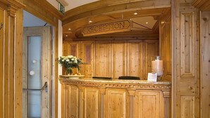 A reception area with a wooden reception desk, a sign above it, and a vase with flowers. - Hotel Garni Alpenstern (Ischgl)