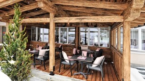 An outdoor wooden deck with tables and chairs, large windows, and a Christmas tree. - Hotel Garni Alpenstern (Ischgl)
