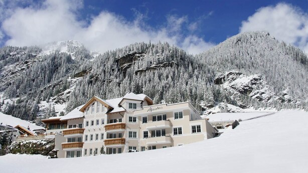 A multi-story hotel with a restaurant on the ground floor, set against a backdrop of snow-covered mountains and trees. - Hotel Garni Alpenstern (Ischgl)