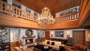 A spacious living room with a fireplace, bookshelves, a chandelier, and a high wooden ceiling. - Hotel Aurelio (Lech am Arlberg)