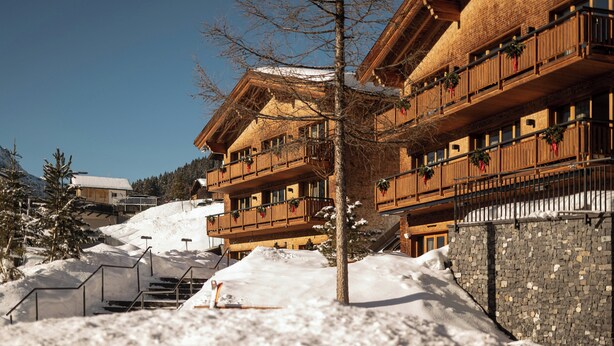A multi-story wooden building with balconies, a stone wall, and a snow-covered slope. - Hotel Aurelio (Lech am Arlberg)