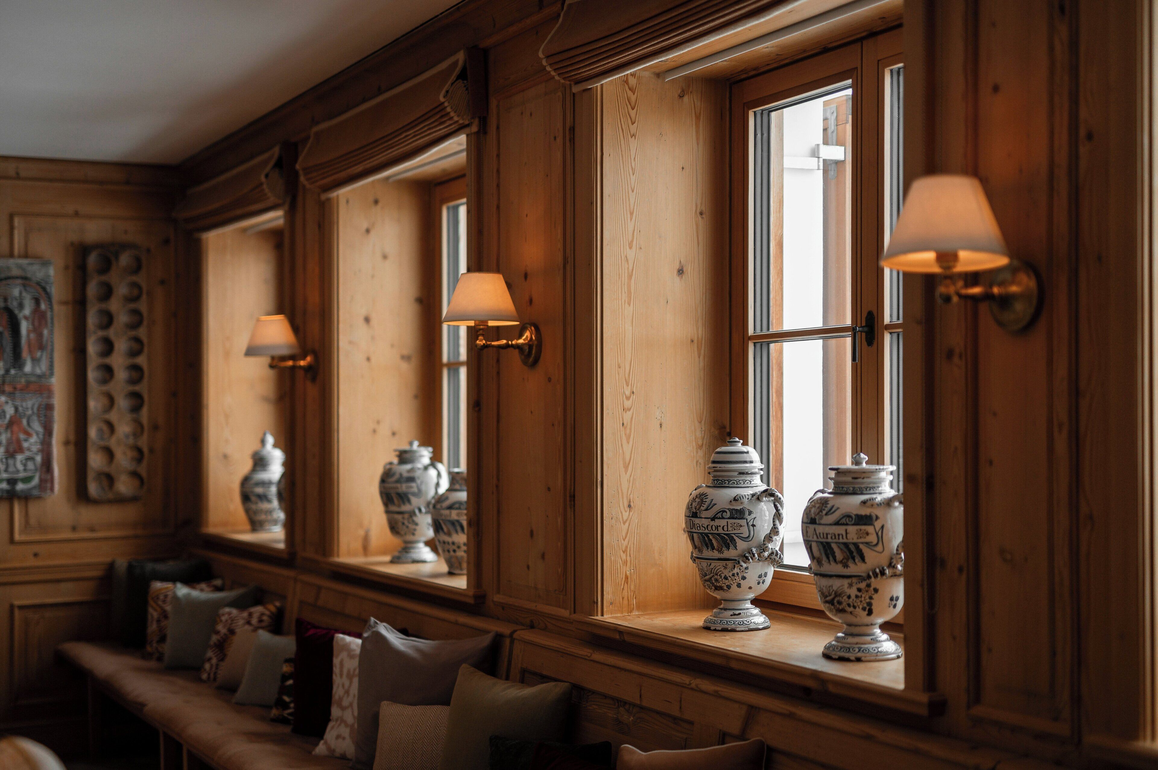 A room with wooden paneled walls, large windows with wall-mounted lamps, and a bench with cushions.