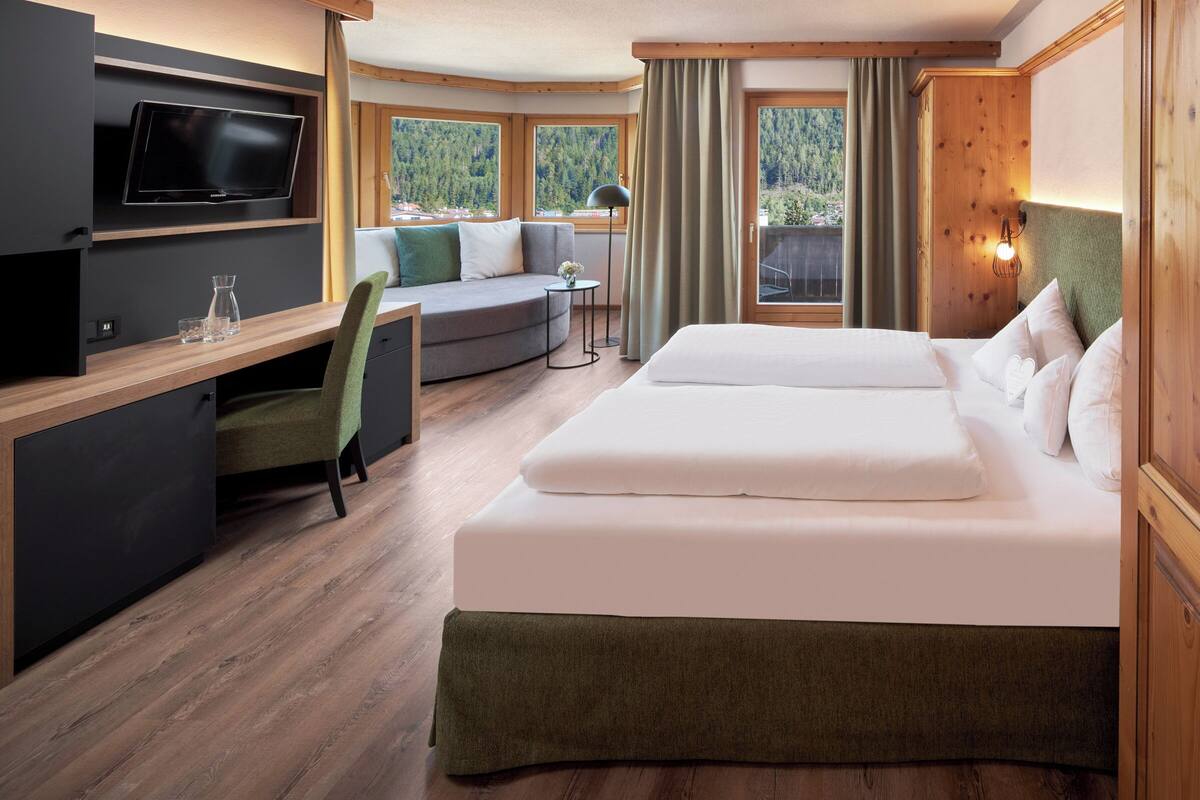 Une chambre d’hôtel moderne équipée d’un lit, d’un bureau avec une télévision, d’un canapé et de grandes fenêtres donnant sur la forêt.