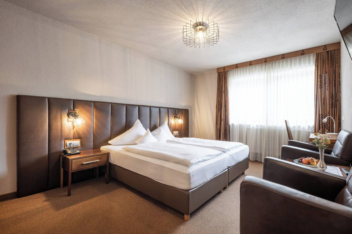 Une chambre d’hôtel avec un grand lit, une table de chevet avec une lampe, une chaise et un bureau agrémenté d’un vase de fleurs.