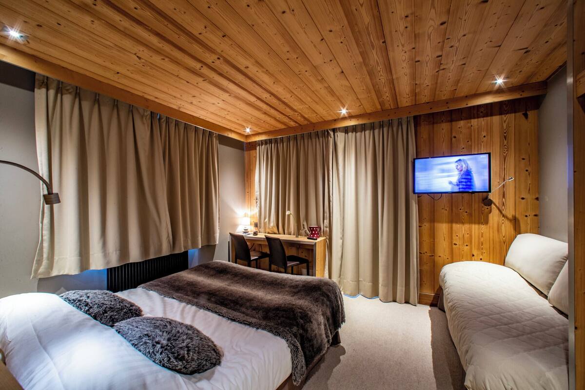 A bedroom with a wooden ceiling, a bed with a fur throw, a desk with a chair, and a TV mounted on the wall.