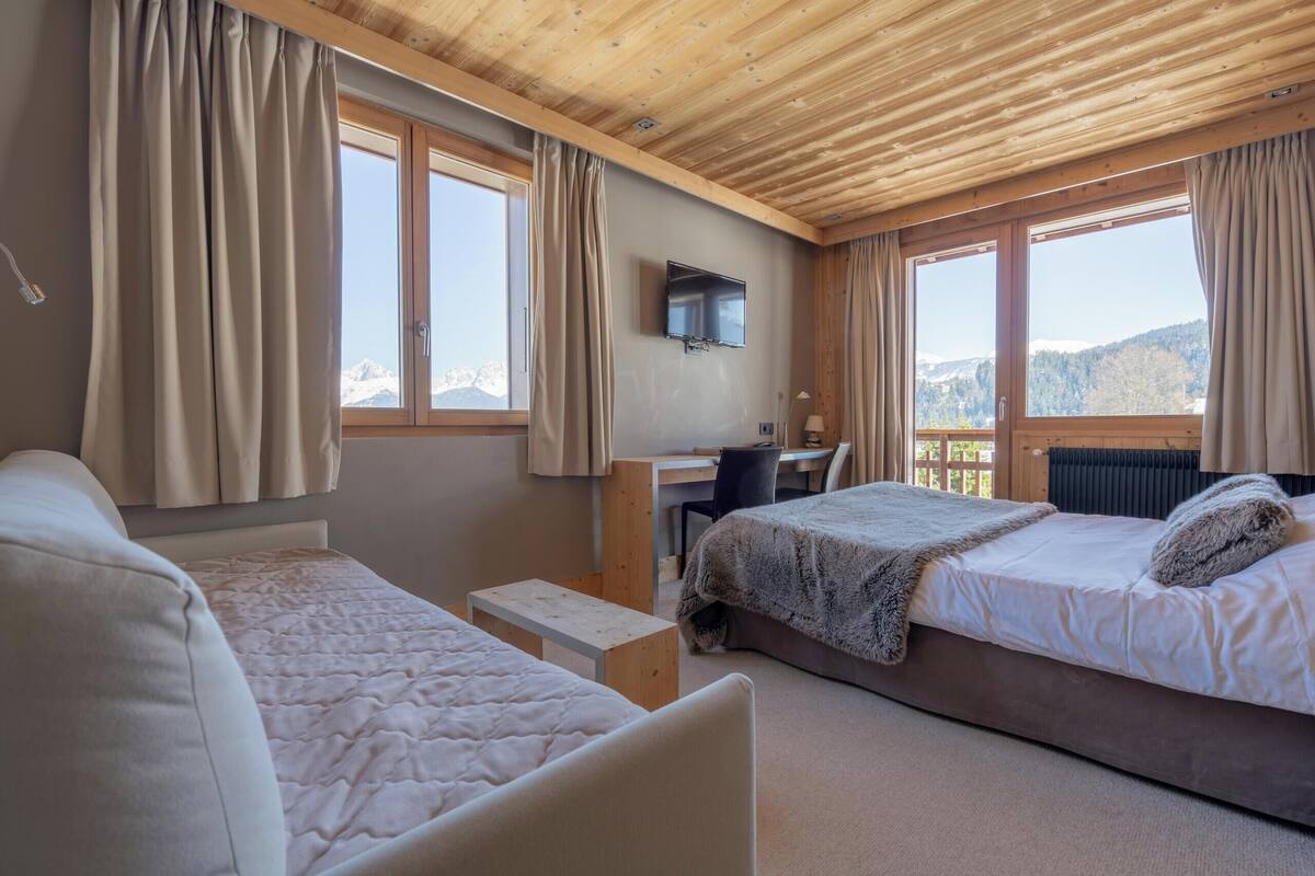 A bedroom with two beds, a desk, a TV, and large windows with curtains.