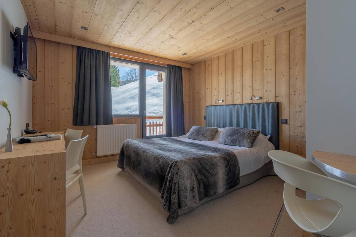 A bedroom with a wooden ceiling, a bed with a dark blanket, a desk with a chair, and a window with curtains.