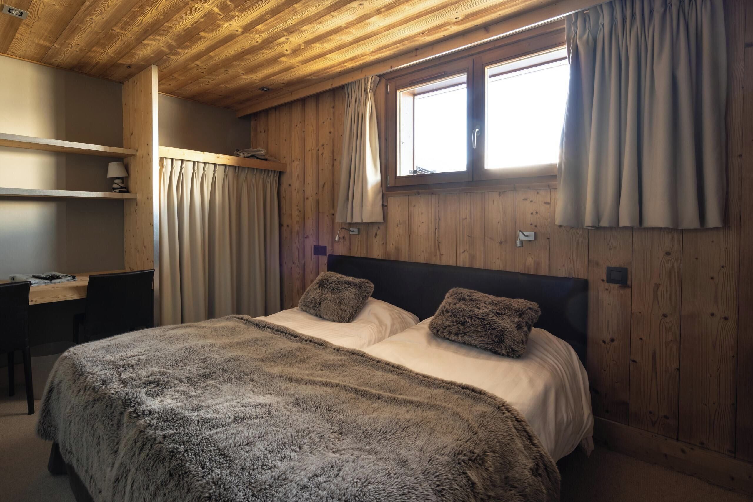 A wooden-paneled bedroom with a bed, two pillows, a desk, and a window with curtains.