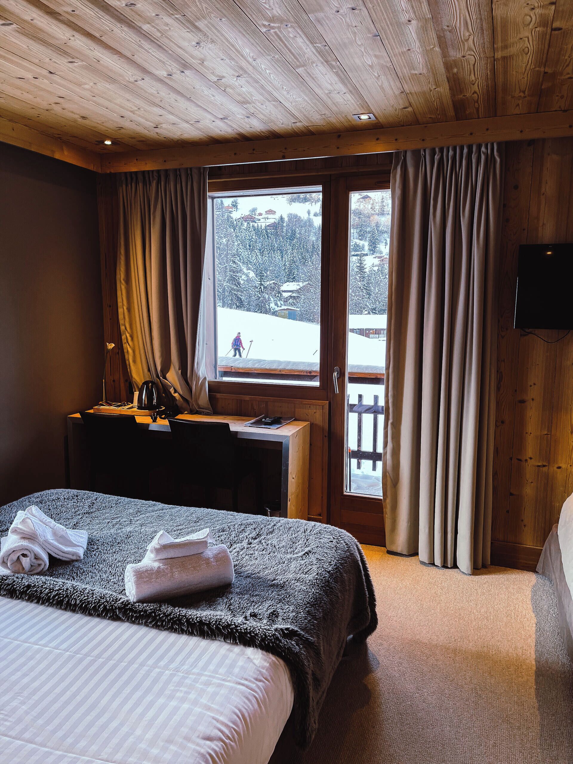 A cozy hotel room with a wooden ceiling, a large bed, a desk with a chair, and a window with curtains.