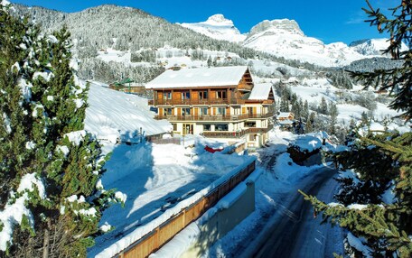 A multi-story wooden chalet with a snow-covered roof, surrounded by snow-covered trees and mountains.