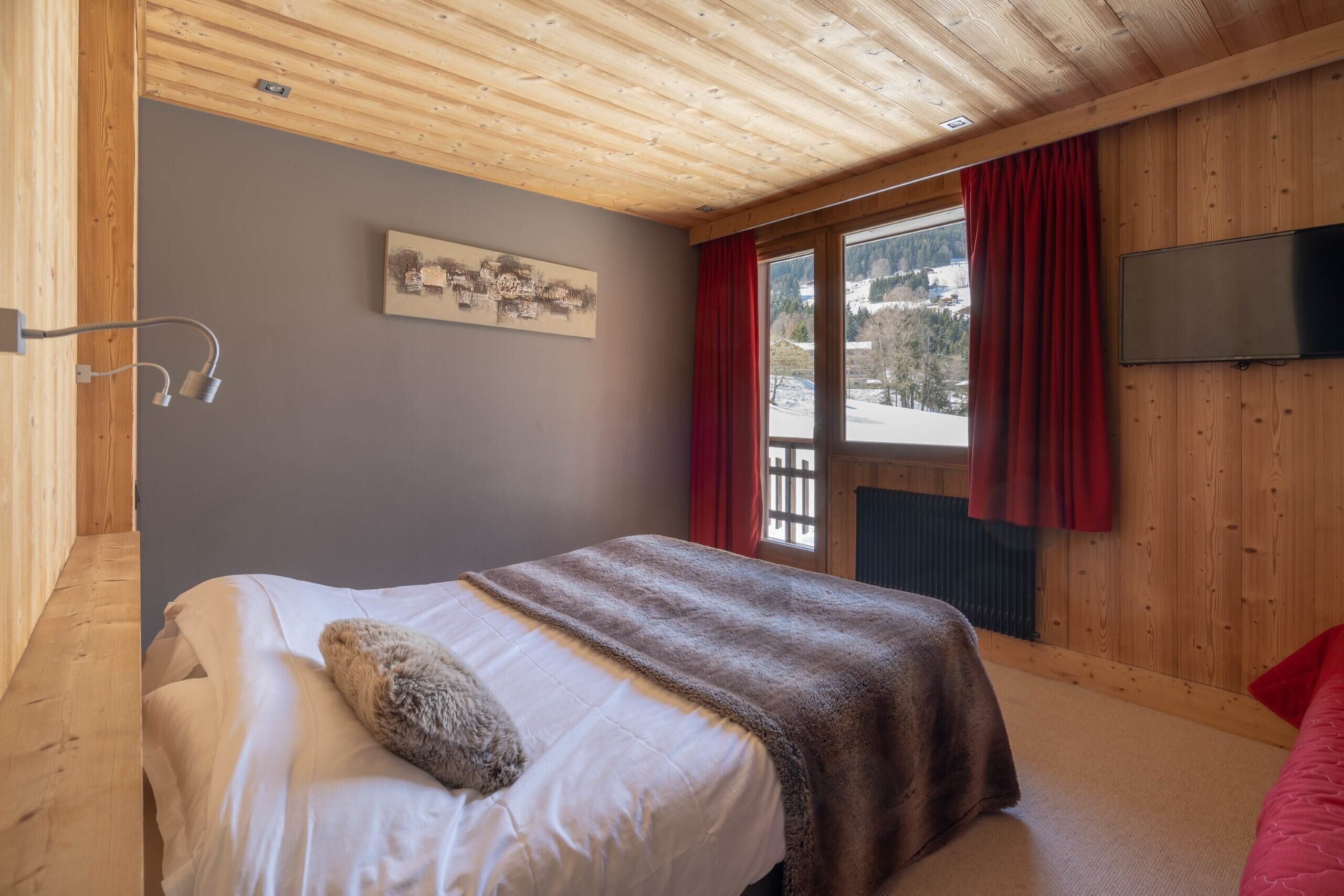 Un dormitorio con techo de madera, cama con colchón blanco y manta marrón, televisor montado y ventana con cortinas rojas.